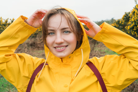 Smiling young woman in a yellow raincoat and hood enjoying walk on background of blooming bush with flowers. Travel concept. Hiking in any weather. Girl on a journey. Relaxing, personal fulfillmentの写真素材