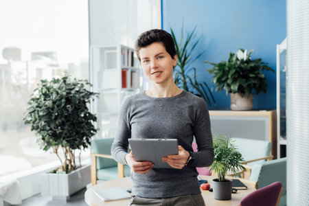 Portrait of smiling middle aged woman in casual outfit with digital tablet standing in company...