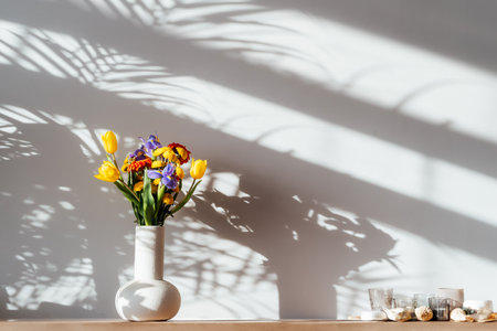 Scandinavian home interior with colorful bouquet of various flowers in ceramic vase standing on wooden cabinet under sunlight and shadows on white gray wall. Minimalist design of home decor.の写真素材