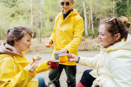 Happy hikers clinking metal mugs, enjoying picnic on bench, drinking hot tea, eating sandwiches in forest. Friends relaxing and having snack picnic on nature background during hiking. Backpacker trip.の写真素材