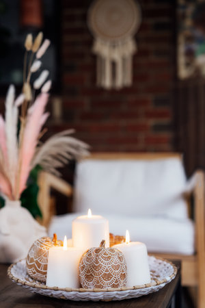 Autumn fall cozy mood composition with natural decor for hygge home. Clay pumpkins decorated with mandalas, white burning candles on wicker tray on coffee table in loft living room.の写真素材