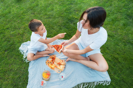 Happy Young mother and preschooler son having outdoor picnic dinner, eating pizza sitting on backyard Lawn on Sunny Day. Happy family time together. Active childhood. House in the suburbs in summer.の写真素材