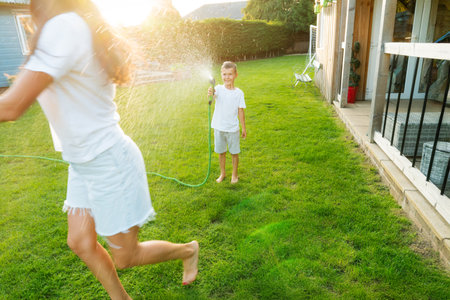 Joyful kid with mother playing with water sprinkler hose, splashing, laughing, having fun together in garden on sunset. Summer outdoor activity. Happy active childhood, family time. Selective focus.の写真素材