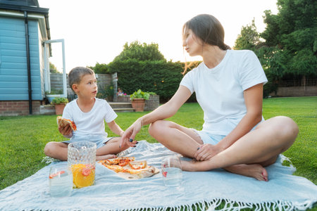 Happy Young mother and preschooler son having outdoor picnic dinner, eating pizza sitting on backyard Lawn on Sunny Day. Happy family time together. Active childhood. House in the suburbs in summer.の写真素材