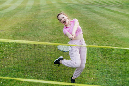 Young shouting focused woman with racket playing badminton outdoors in urban city park. Friends...