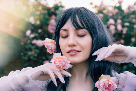 Fairy tale woman portrait. Young beautiful woman Fairy holds pink flower rose on blooming bush background. Hairstyle loose green hair, romantic purple medieval vintage dress. Summer fantasy garden.の写真素材