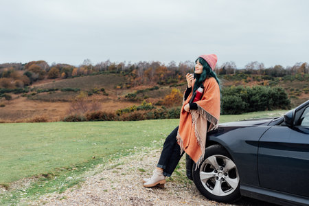 Hipster Woman sits on cars hood, wrapped in warm Shawl Wraps Poncho Cape and hat and enjoying warm drink, while overlooking rural landscape during road trip. Cold season travel, vacation conceptの写真素材