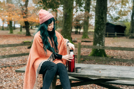 Woman wrapped in Shawl Poncho sits on picnic table pouring hot drink from thermos during autumn walk in forest with fall leaves. Feeling harmony, relax, personal fulfillment. Local travel lifestyle.の写真素材