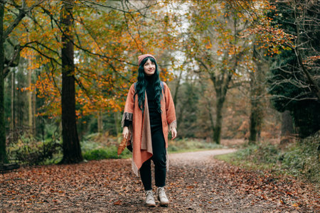 Hipster woman wrapped in Shawl Poncho with backpack enjoying autumn walk in forest with fall leaves. Feeling harmony, reunion with nature. Relaxing, personal fulfillment. Local travel lifestyle.の写真素材