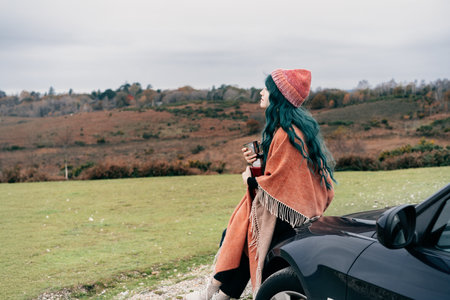 Hipster Woman sits on cars hood, wrapped in warm Shawl Wraps Poncho Cape and hat and enjoying warm drink, while overlooking rural landscape during road trip. Cold season travel, vacation conceptの写真素材