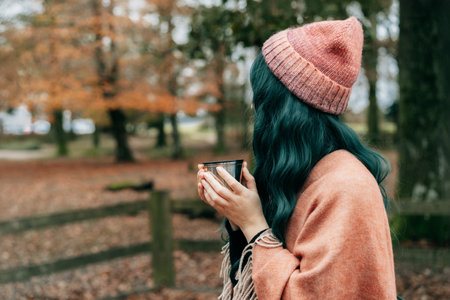 Side view Woman in hat wrapped in Shawl Poncho drinking hot drink from thermos cup during autumn walk in forest with fall leaves. Feeling harmony, relax, personal fulfillment. Local travel lifestyle.の写真素材