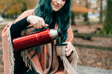 Woman wrapped in Shawl Poncho sits on picnic table pouring hot drink from thermos during autumn walk in forest with fall leaves. Feeling harmony, relax, personal fulfillment. Local travel lifestyle.の写真素材