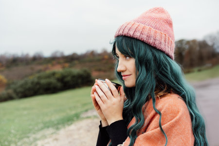 Hipster Woman sits on cars hood, wrapped in warm Shawl Wraps Poncho Cape and hat and enjoying warm drink, while overlooking rural landscape during road trip. Cold season travel, vacation conceptの写真素材
