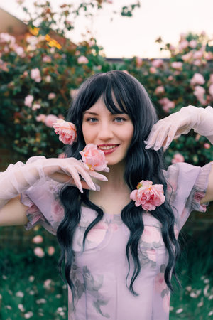 Fairy tale woman portrait. Young beautiful woman Fairy holds pink flower rose on blooming bush background. Hairstyle loose green hair, romantic purple medieval vintage dress. Summer fantasy garden.の写真素材
