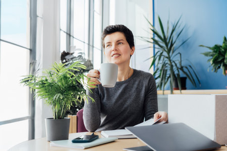 Satisfied smiling newcomer middle aged neutral gender female employee holds coffee cup and sitting at her new office desk work space. First working day concept. Dreaming about new opportunitiesの写真素材