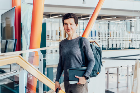 Smiling neutral gender middle-aged female person in casual clothes with backpack and books stands on staircase in modern building of university or education Centre. Adult students, Academic education.の写真素材