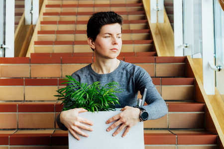 Stressed worried middle aged gender neutral woman with box of personal items sitting alone on the staircase after being fired, lost job due to recession and economic stress. Equality discrimination.の写真素材