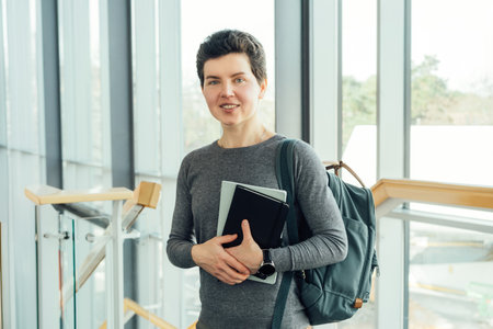 Smiling neutral gender middle-aged female person in casual clothes with backpack and books stands on staircase in modern building of university or education Centre. Adult students, Academic education.の写真素材