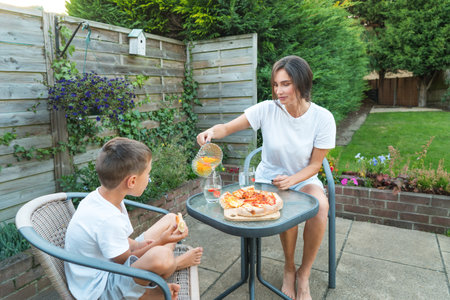 Happy Young mother and son having fresh air dinner, eating pizza with lemonade while sitting on patio outdoor furniture. Happy family time together. Active childhood. House in the suburbs in summerの写真素材