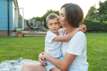 Happy Young mother kissing her cute Caucasian preschooler kid who embracing. Young mom and kid hugging with love, affection, tenderness on the backyard lawn background on sunset. Time together.の写真素材