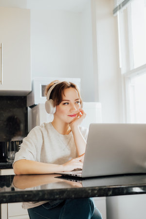 Dreaming young Woman in headset enjoying and listen to music while working on laptop at Kitchen Table in tiny flat. Take a break for recovery, mental health. Relax, no stress concept. Vertical card.の写真素材
