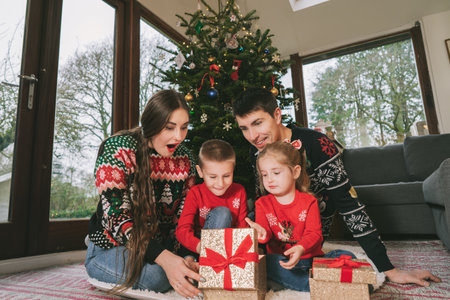 Happy family in festive sweaters sitting under Christmas tree at home. Emotional parents and kids feeling excited unwrapping Christmas gifts, having fun, Family time, childhood miracle time concept.の写真素材