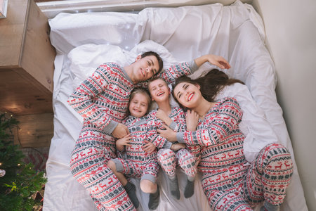 Top view happy family in festive pajamas playing on bed on Christmas Eve in their apartment. Dad, mom,daughter, son are lying on bed and having fun together. Family celebrating winter holidays at homeの写真素材