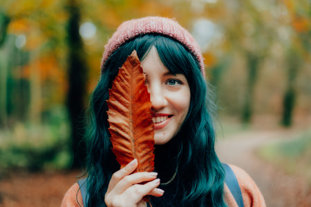 Portrait of Young smiling woman in hat partially covering her face with striking red fallen leaf enjoying autumn walk in forest. Feeling harmony, reunion with nature. Relaxing, personal fulfilmentの写真素材