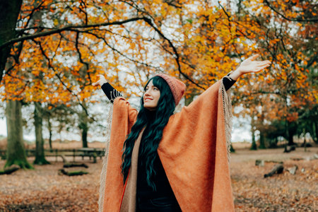 Smiling woman in hat wrapped in warm Shawl Poncho raised arms enjoying life and freedom in autumn...
