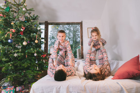 Happy family in festive pajamas playing on bed on Christmas Eve in decorated apartment. Dad, mom,daughter, son are lying on bed and having fun together. Family celebrating winter holidays at homeの写真素材