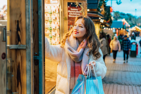 Happy woman enjoys shopping at festive Christmas market while carrying shopping paper bags, walking in street Xmas fair of European city. Selecting gifts for New Year, winter holiday sales, discountsの写真素材