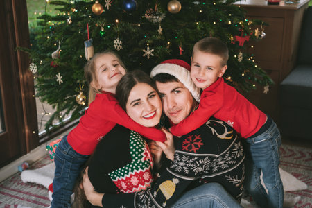 Smiling family portrait in festive sweaters sitting with gifts under Christmas tree at home during holiday. Happy parents and kids cuddling, feeling excited. Family time together. Happy childhoodの写真素材