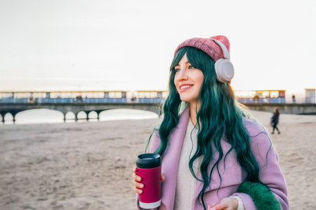 Stylish young smiling hipster woman with color hair wearing wireless headphones, pink coat, hat, walking on beach with reusable coffee cup. Time To Relax. seasonal fashion style trend. Barbiecore.の写真素材