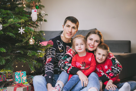 Smiling family portrait in festive sweaters sitting with gifts under Christmas tree at home during holiday. Happy parents and kids cuddling, feeling excited. Family time together. Happy childhoodの写真素材