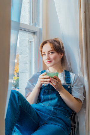 Free Time To Relax at home. Portrait of young woman siting on the windowsill at cozy home holds cup of hot tea drink. Happy calm female taking break for mental health wellbeing. Vertical card.の写真素材