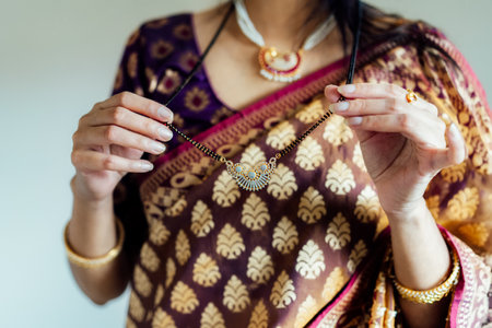 Dressed in beautiful saree, Indian woman delicately putting traditional jewellery necklace getting ready for ethnic festive celebration. Indian culture, ritual holiday in modern life. Selective focus.の写真素材