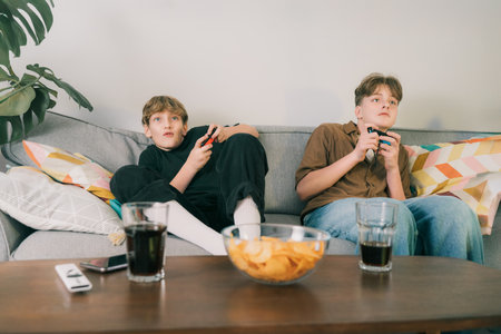 Two teenager boys are focused on their video game consoles while sitting on couch at home. Brothers...