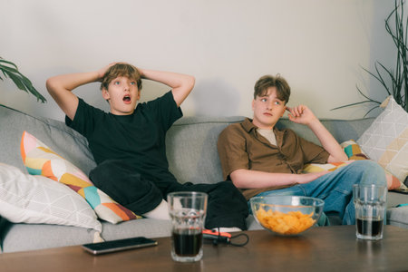 Two excited teenager boys watching match game together at home. Brothers seated on couch with snacks and drinks, cheering for favourite team but are Disappointed after Team Loses. Young sport fansの写真素材