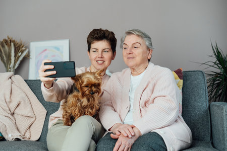 Neutral gender woman hugging her senior mom or grandma and taking a selfie by phone with a small dog nestled in womans lap, creating warm family moment. Parenting love, care and generations unityの写真素材