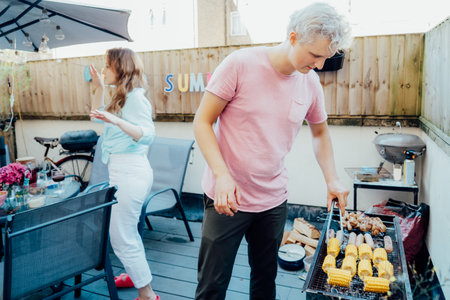 Friends enjoying summer party on private outdoor patio terrace with drinks and snacks. Friends celebrate summer on bright patio, with male grilling BBQ and female playfully dancing aroundの写真素材