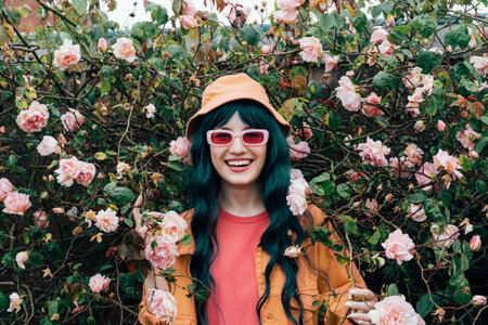 Young laughing hipster woman in bright outfit, bucket hat and sunglasses posing on blooming roses...