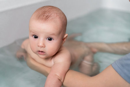 Cute baby bathing and swimming in the bath. Baby learning to swim and playing sports. Father enjoys bath time with his curious baby in a cozy bathroom. Fatherhood concept. Selective focus.の写真素材