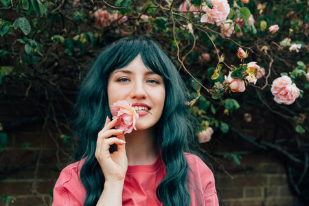 Romantic young woman with long green hair smiles while holding a delicate rose in her mouth amidst beautiful blooming flowers in a vibrant garden. Spring or Summertime vibes, fun, positive mood.の写真素材