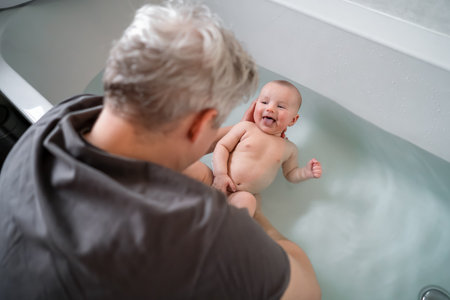 Top view smiling baby bathing and swimming in the bath. Baby learning to swim and playing sports. Father enjoys bath time with his curious baby in a cozy bathroom. Fatherhood concept. Selective focus.の写真素材