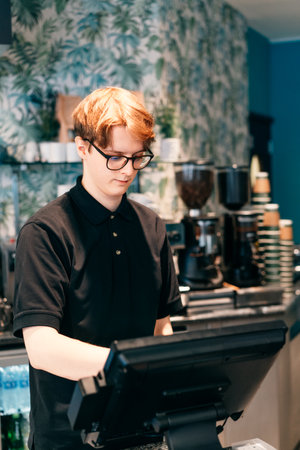 Young waiter serving customer at cash point in cafe. Man working with POS terminal. Cashier, barista checking for payment receipt. Hospitality, server and preparing a slip at the till in coffee shopの写真素材