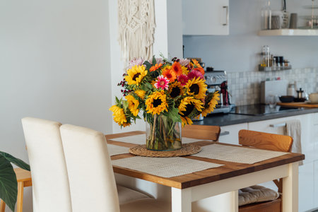Stylish modern interior of open space white kitchen with huge multicolor summer flower bouquet in vase on wooden kitchen counter table, macrame on the wall. Cozy Design home decor. Selective focus.の写真素材