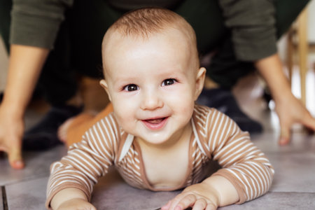 Happy, laughing baby lying on tiled floor at home with no face father on background. Dad And Toddler Child Enjoying Spending Time Together, Cheerful active infant smiling, learning to crawl, playingの写真素材