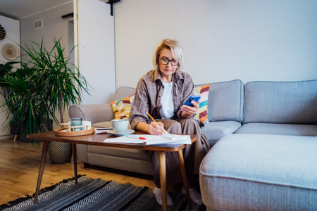 Middle aged woman reviewing expenses, managing personal finances on smartphone mobile app while sitting on couch with coffee in cozy living space. finance planning, financial balance check, budgetingの写真素材