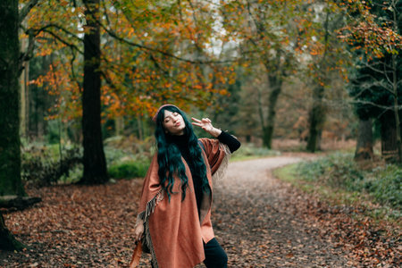 Young hipster Woman wrapped in warm Shawl Poncho grimacing, making V sign by fingers during autumn walk in forest with fall yellow leaves. Emotional expression. Travel lifestyle. Relaxing, having fun.の写真素材