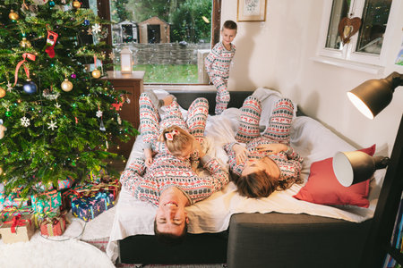 Happy family in festive pajamas playing on bed on Christmas Eve in decorated apartment. Dad, mom,daughter, son are lying on bed and having fun together. Family celebrating winter holidays at homeの写真素材
